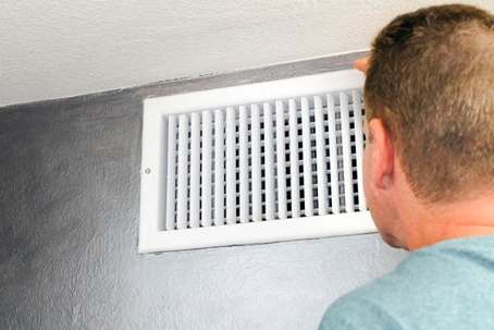 A man examines a white wall vent in the ceiling.
