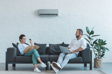 A man and woman relax on a couch, using a laptop and a remote, below a wall-mounted air conditioner.