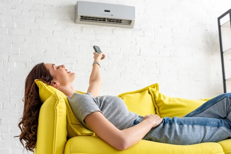 A woman reclines on a yellow couch, pointing a remote at a wall-mounted air conditioner.