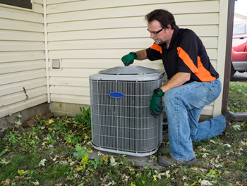 A man in work clothes inspects a home air conditioning unit outside.