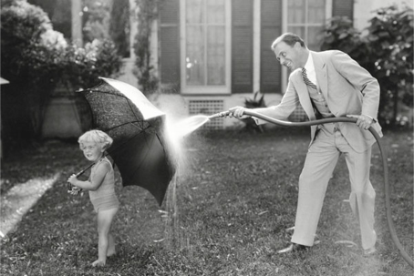 A black and white photo shows a man spraying a hose toward a small child holding an umbrella.